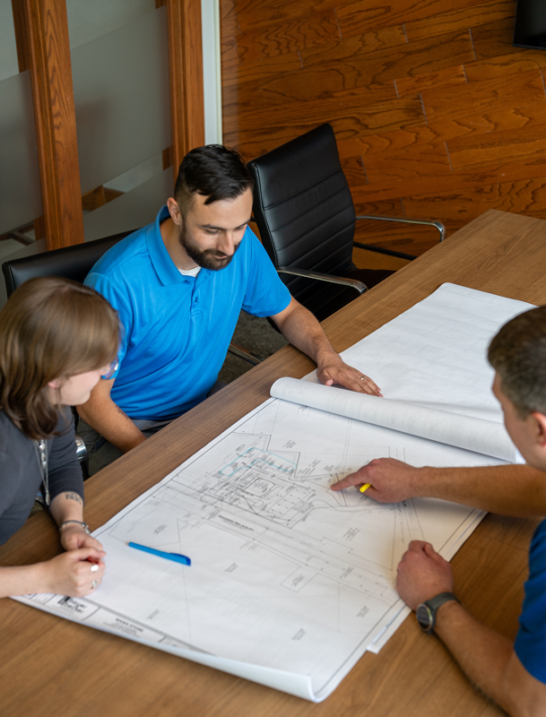 Three people reviewing blueprints at a wooden table.
