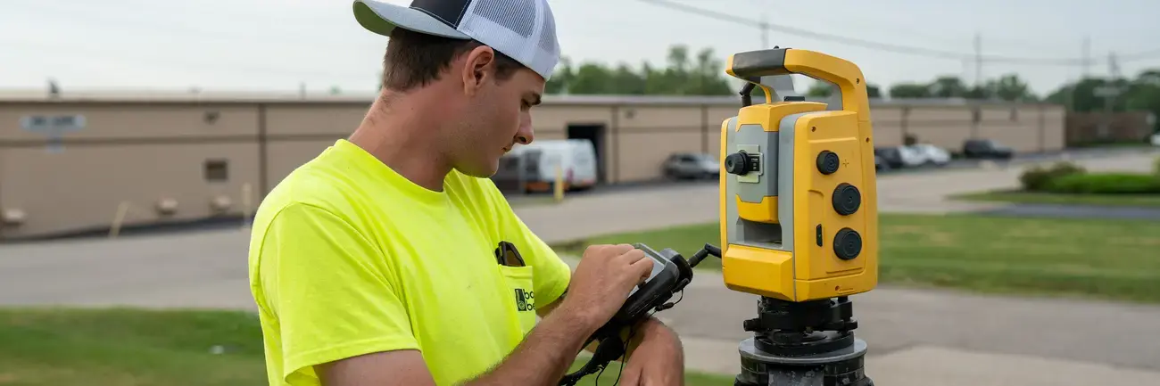 Bayer Becker Surveying Man in yellow shirt adjusting survey equipment on tripod outdoors.